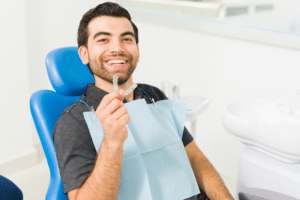 Smiling man sitting in a blue dental chair holding up a clear aligner tray.