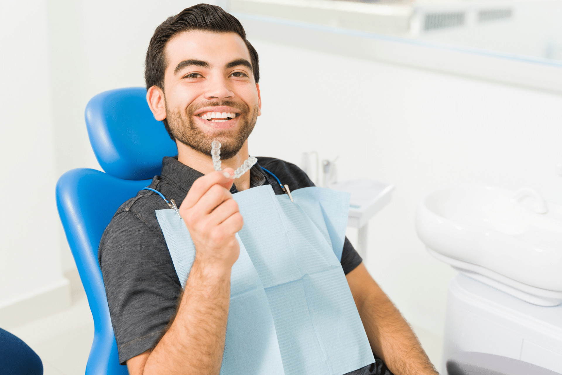 Smiling man sitting in a blue dental chair holding up a clear aligner tray.